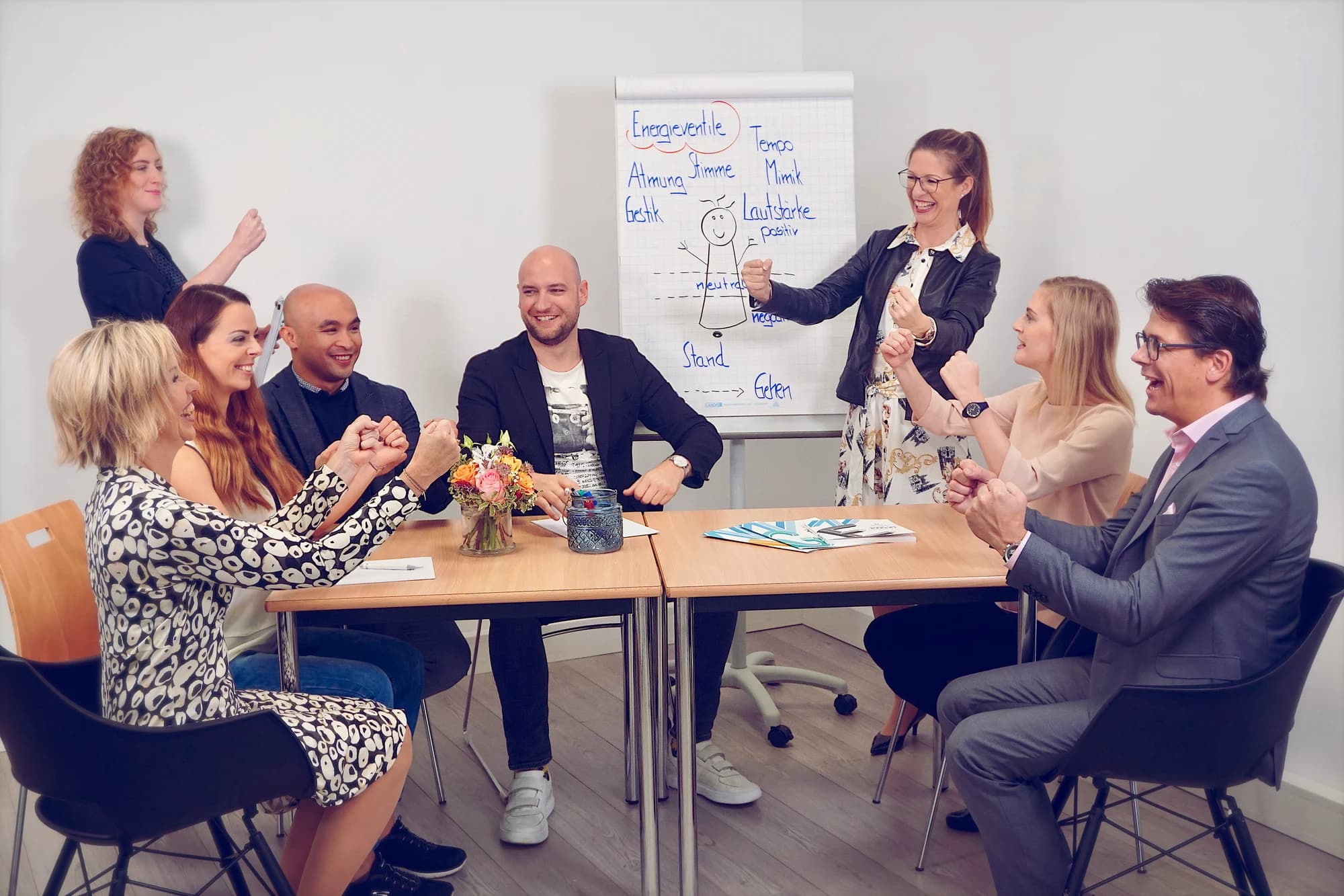 Seminargruppe an einem Tisch, davor eine Trainerin an einem Flipchart